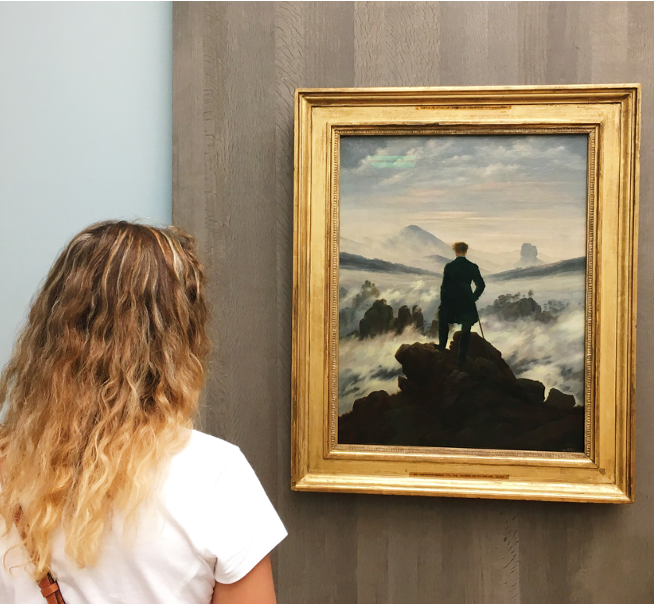 A woman with wavy, blonde hair, wearing a white shirt, stands in front of a framed painting in a museum. As a TCK, she is captivated by the scene that depicts a lone figure standing on a rocky outcrop, gazing out over a misty landscape with mountains in the distance.