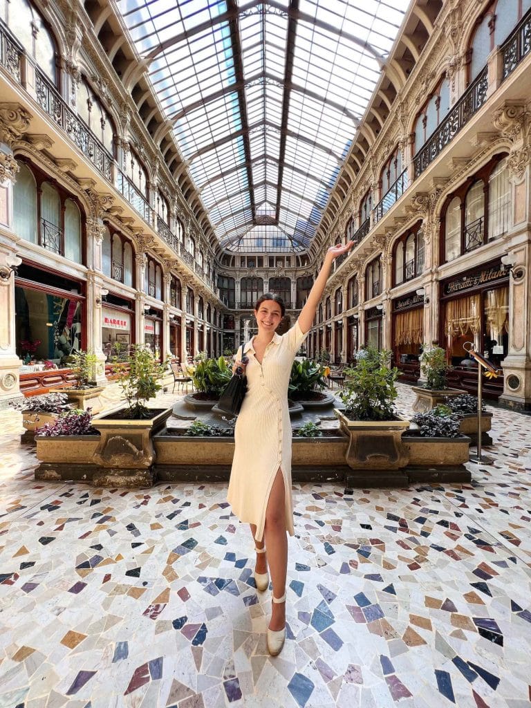 A TCK woman in a cream-colored dress stands in the center of a grand indoor gallery with a glass ceiling. She is smiling, one arm raised as if waving. The gallery, Galleria Subalpina, has ornate architecture with balconies, plants, and decorative tiles on the floor.