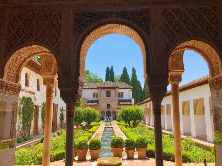 A scenic view through ornate arches reveals a lush garden with a central water feature, leading to a historic building. Exploring such serene courtyards is one of the best things to do in Granada, where tall cypress trees and a clear blue sky frame the picturesque scene.