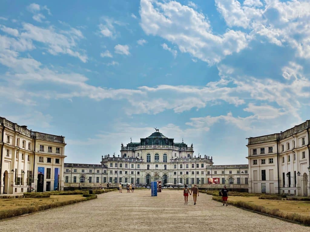 A grand historic palace, Palazzina di Caccia di Stupinigi, with a symmetrical facade and domed roof stands under a partly cloudy blue sky, its wide gravel courtyard filled with visitors—one of the best things to do in Turin for history and architecture lovers.