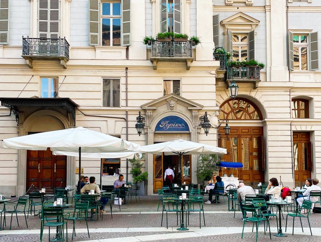 Outdoor café scene in front of an elegant European building with tall windows and balconies, reminiscent of Turin historic cafes. Green chairs under white umbrellas seat several people at tables, enjoying the day.