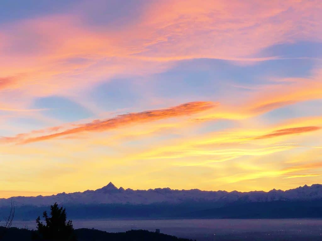 Colorful sunrise sky with shades of pink, orange, and yellow above a silhouette of distant Italian Alps and trees. These are the views from Superga, Turin's tallest hill - a must visit!