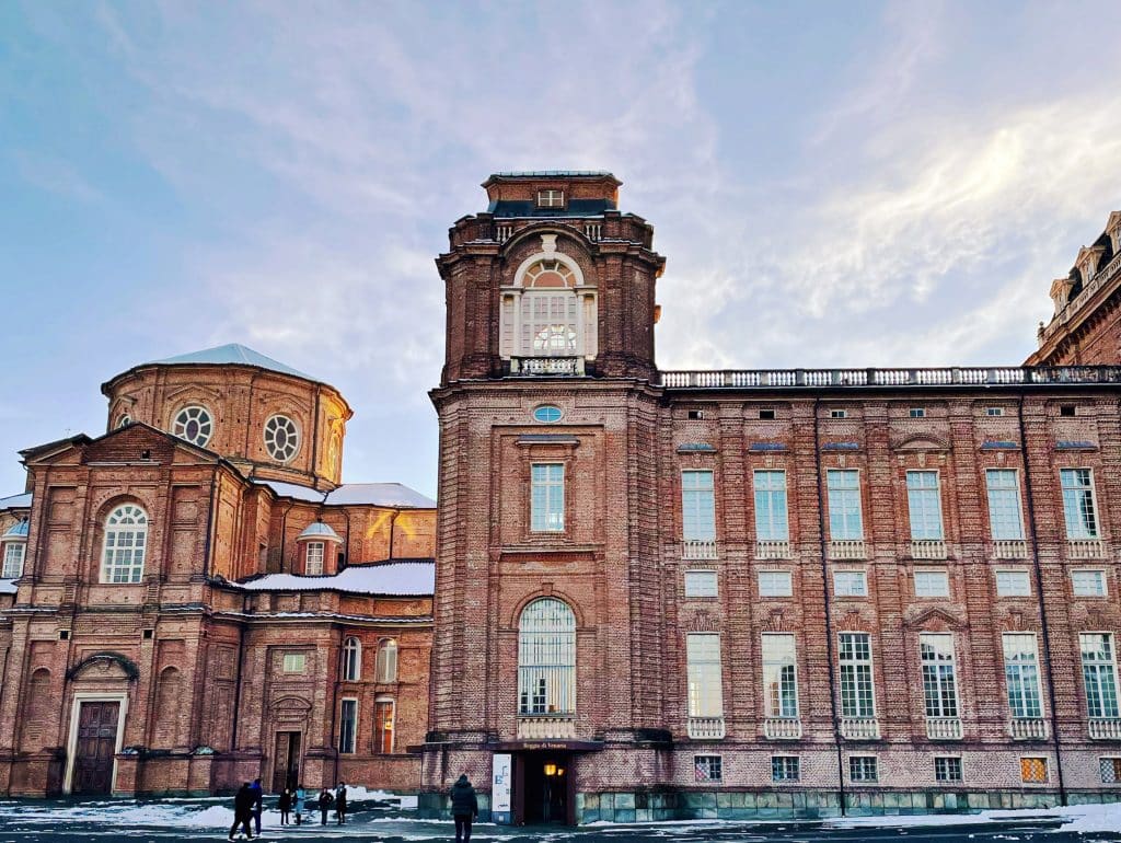 A large historic brick palace, La Venaria Reale, with tall windows and ornate architecture, bathed in warm sunset light, is a must-see when in Turin.