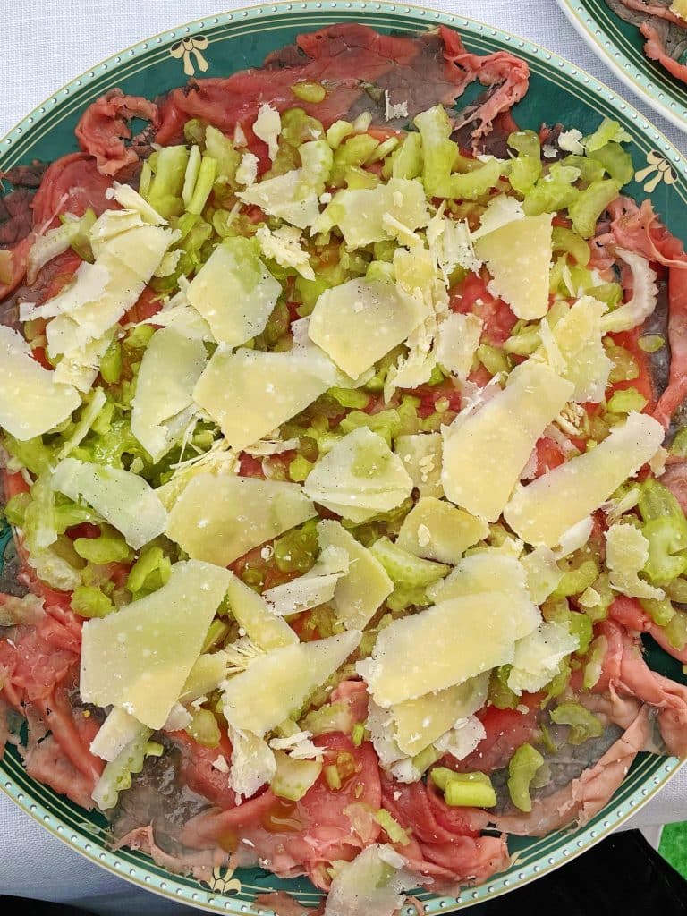 A plate of carpaccio topped with thinly sliced raw beef, celery, and large Parmesan shavings, beautifully arranged on a green floral dish—just one of many reasons to visit Turin and experience its exquisite culinary scene.