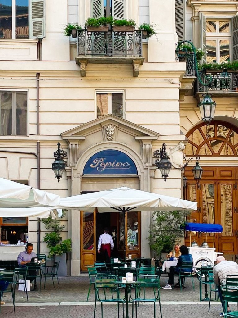 Outdoor historic café scene with green chairs and tables, people dining under white umbrellas, and a waiter entering a building labeled "Pepino." Charming balconies and potted plants hint at reasons to visit Turin—this vibrant city is worth visiting for its lively street life.