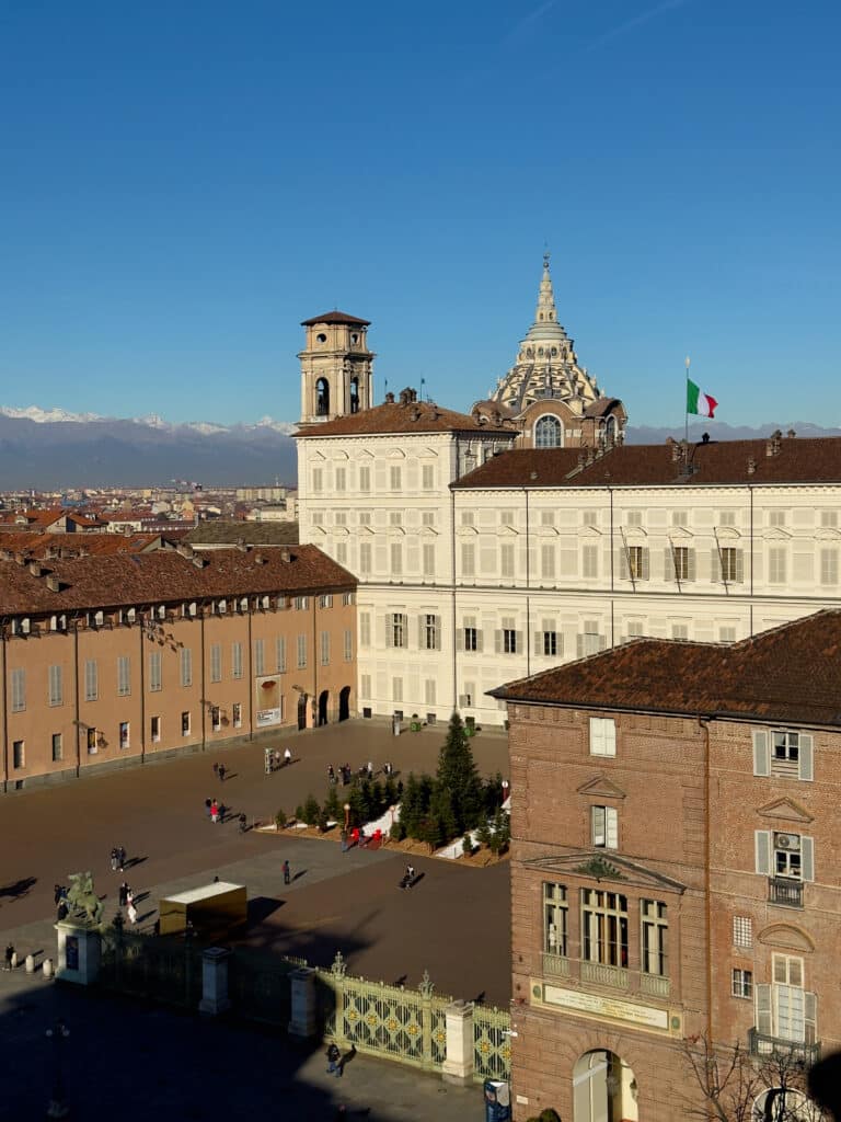 A sunny view of Piazza Castello in Turin, Italy, featuring the white Palazzo Reale—one of the Royal Residences of the House of Savoy—a must visit in Turin, with the Italian flag, distant mountains, and people walking in the square.
