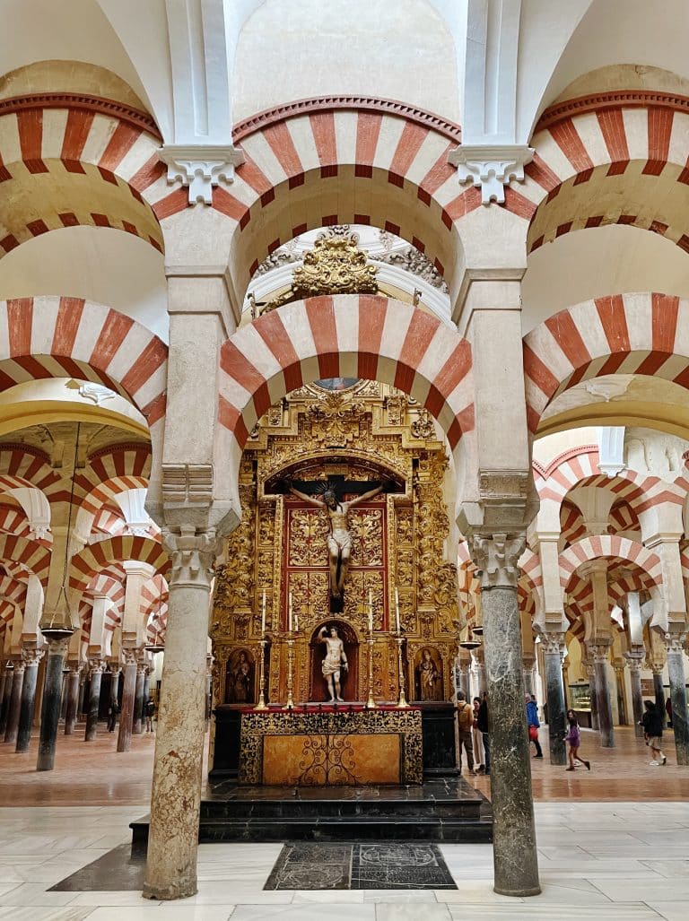 A richly decorated golden altar with a crucifix stands beneath red-and-white striped arches in the Mezquita-Catedral of Córdoba, Spain—a stunning highlight often included in day trips from Granada. Marble columns support the arches as visitors explore.