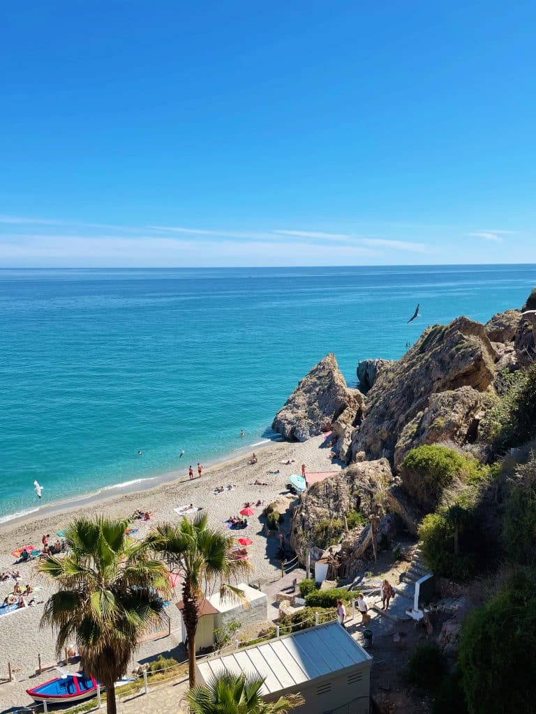 A sunny beach scene in Nerja with turquoise water, people sunbathing and swimming, rocky cliffs, palm trees, and a clear blue sky—perfect for day trips from Granada.