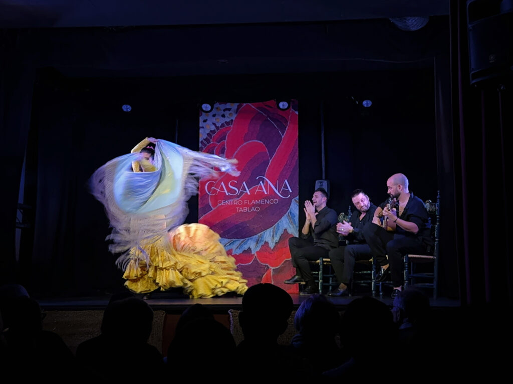 A flamenco dancer in a yellow dress twirls onstage, her shawl flowing, while three musicians sit to the right, clapping and playing guitar. The backdrop reads "Casa Ana Centro Flamenco Tablao" with colorful abstract art.