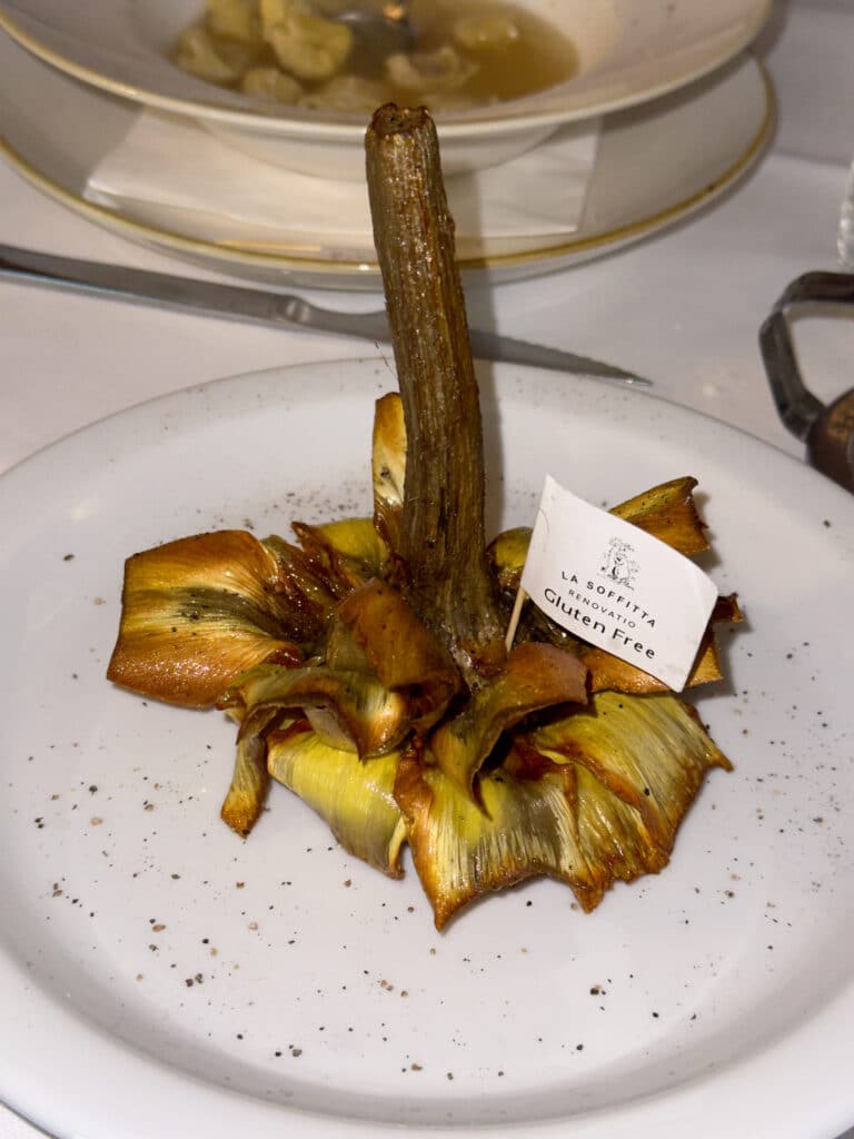 A plated fried artichoke sits upright with crisp, golden petals spread out, seasoned with pepper. A small tag reading "Gluten Free" is placed on top. In the background, another dish is partially visible.