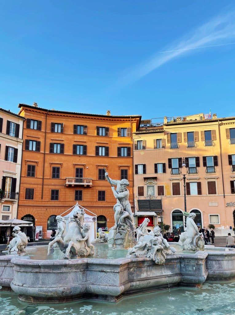 A white marble fountain with intricate statues of people and horses stands in a plaza, surrounded by colorful historic buildings under a clear blue sky—a must-see during your 3 days in Rome itinerary.
