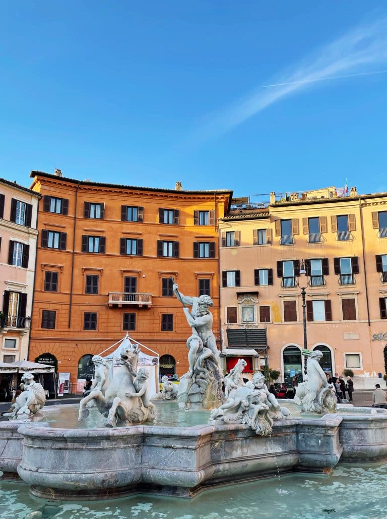 A white marble fountain with intricate statues of people and horses stands in a plaza, surrounded by colorful historic buildings under a clear blue sky—a must-see during your 3 days in Rome itinerary.