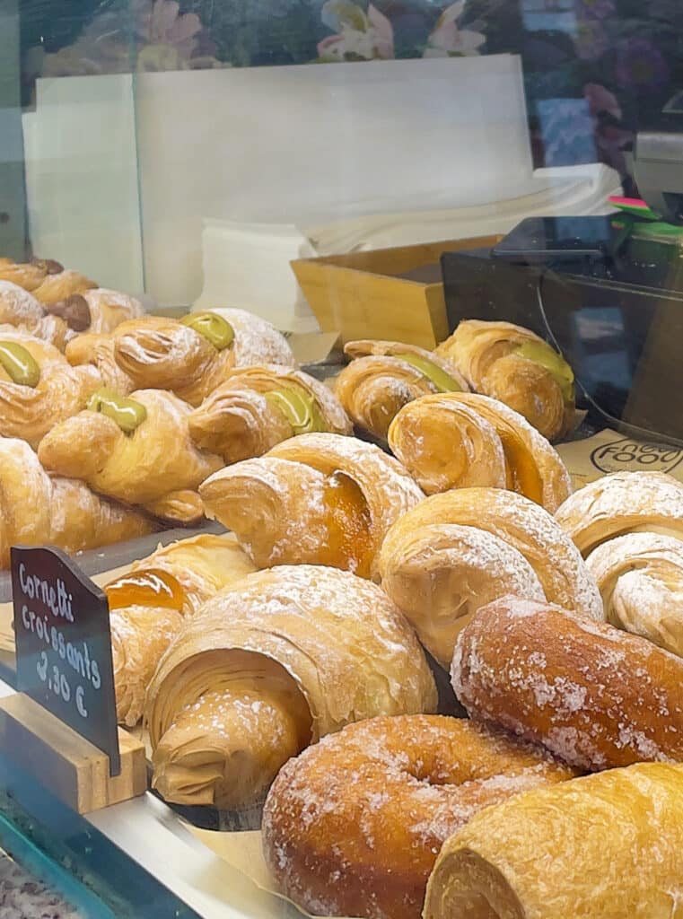 A display case filled with various pastries, including croissants dusted with powdered sugar and sugar-coated donuts, alongside a small sign reading "Cornetti croissants €1.30"—a tempting find for anyone seeking gluten free Rome treats.