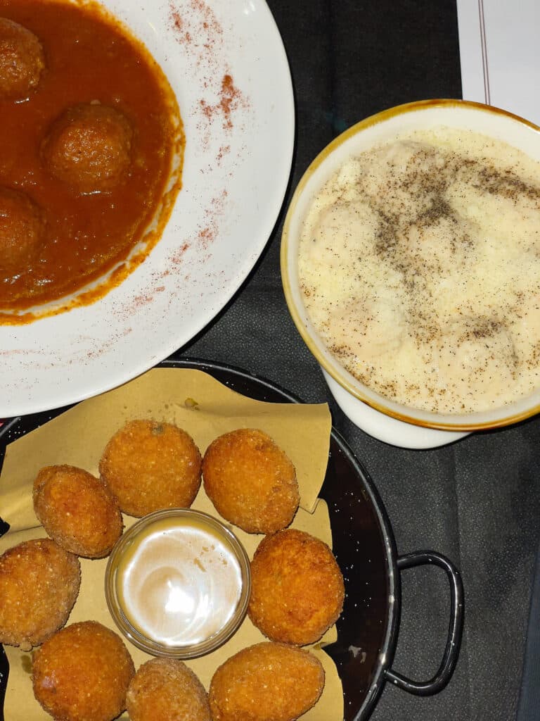 Three dishes on a dark table: meatballs in tomato sauce, a bowl of pasta topped with grated cheese and pepper, and gluten free Rome-inspired round breaded appetizers with a dipping sauce in the center.