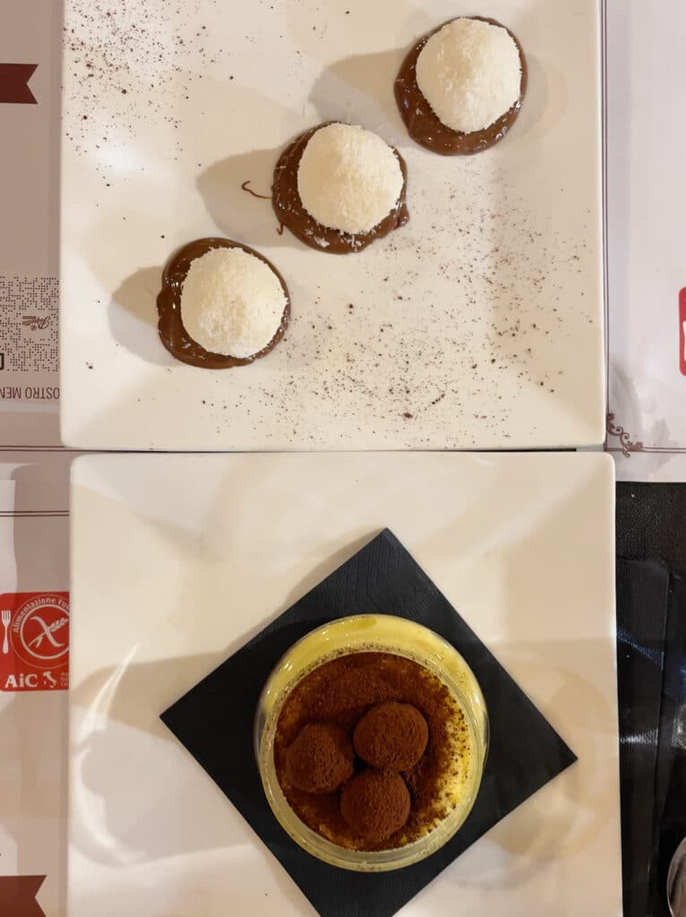 Two square white plates: the top plate has three round chocolate and coconut desserts, while the bottom holds a glass dish of gluten free tiramisu topped with cocoa-dusted truffles—an indulgence you can enjoy in Rome.