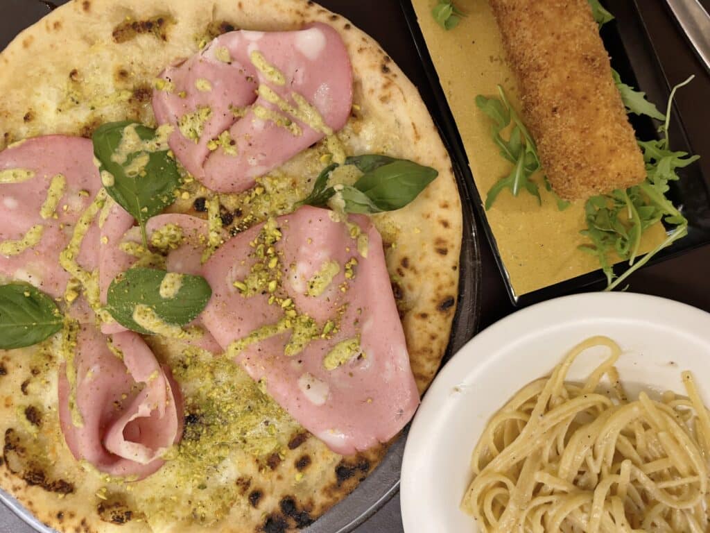A close-up of a pizza topped with mortadella, basil, and crushed pistachios, next to a plate of creamy pasta and a rectangular breaded fried appetizer on arugula—perfect for sampling gluten free Rome delights.