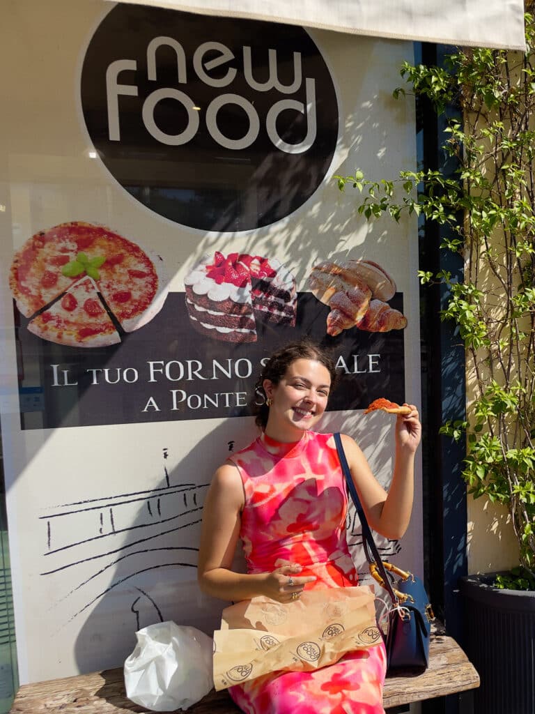 A woman in a pink and red dress sits on a bench outside a bakery in Rome, smiling and holding a gluten free pizza al taglio. Behind her is a sign with images of food and the words "new food" and "IL TUO FORNO ARTIGIANALE A PONTE S.