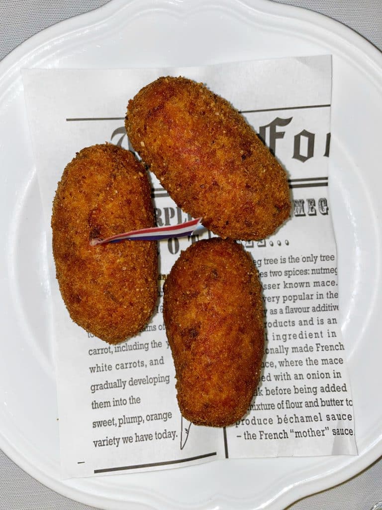 Three golden-brown croquettes, perfect for a gluten free Rome experience, are arranged on a white plate lined with printed paper. One croquette has a toothpick inserted. The plate rests on a white background.
