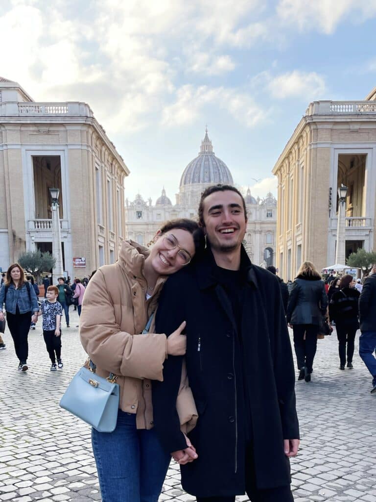 A smiling couple stands close together in a plaza, with the woman leaning her head on the man’s shoulder. They are surrounded by people, and St. Peter’s Basilica is visible in the background under a blue sky with clouds.