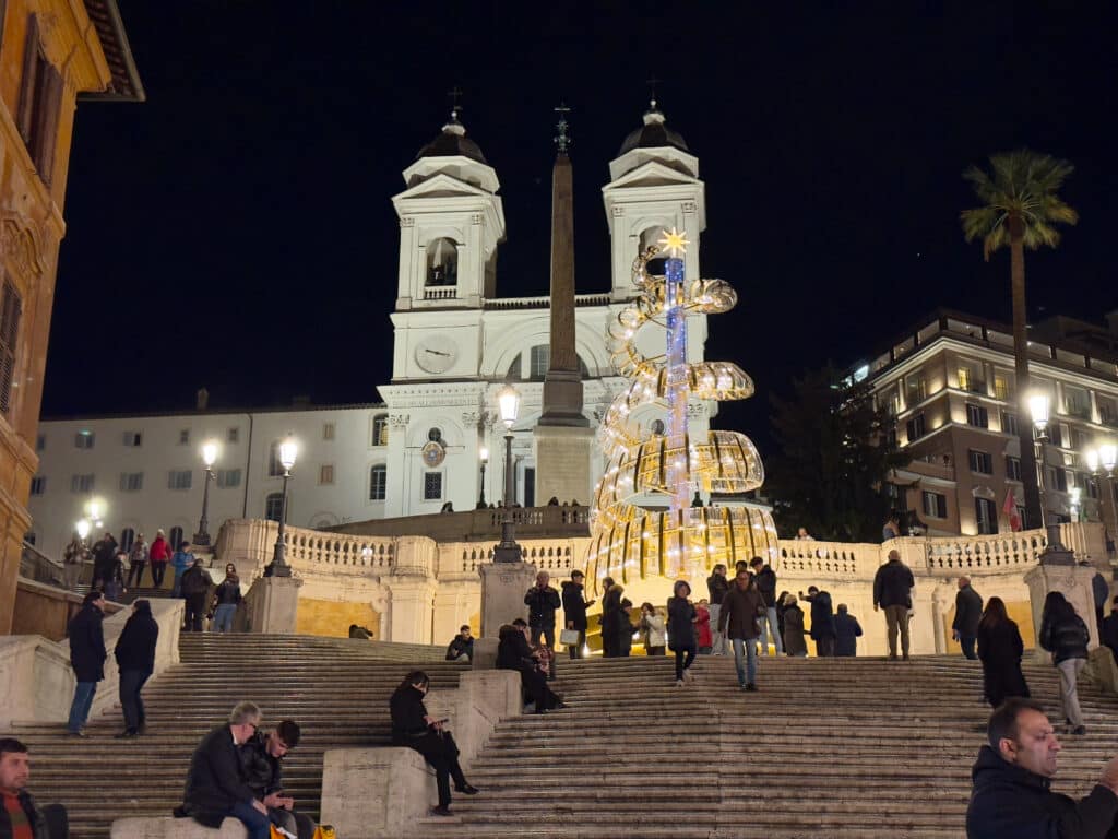 People gather on the Spanish Steps in Rome at night, with the Trinità dei Monti church in the background and a modern, illuminated Christmas tree sculpture in the center of the steps.