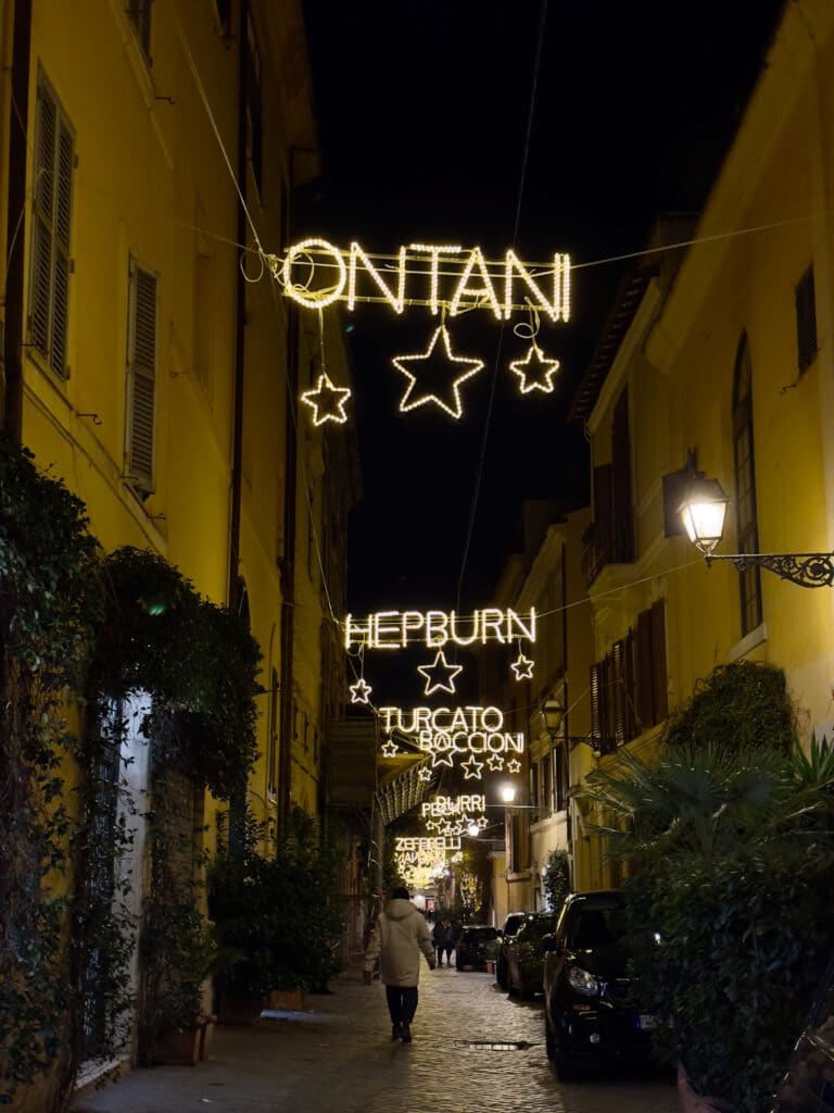A narrow, cobblestone street at night is decorated with hanging illuminated signs of names and stars. Buildings line both sides, and a person walks alone under the glowing lights.