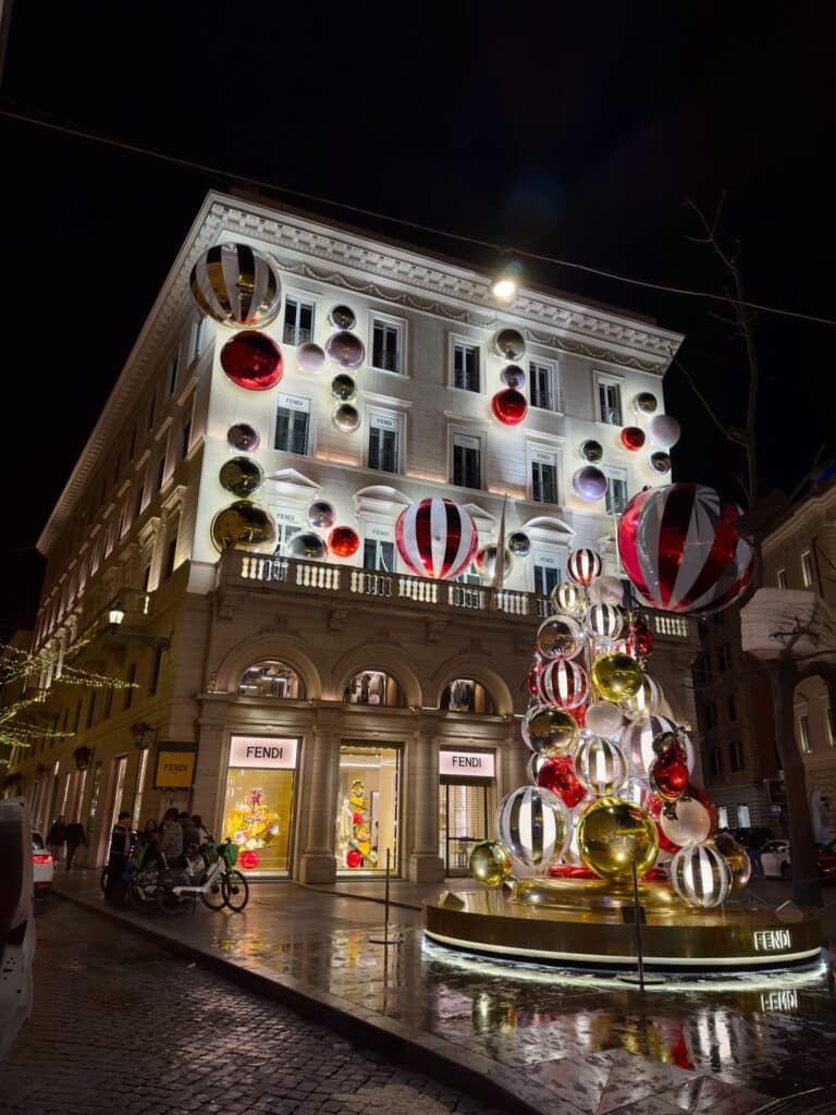 A Fendi store on a city street at night, decorated with large, colorful holiday ornaments on the building facade and a display in front. The building is brightly lit, reflecting on the wet cobblestone street.