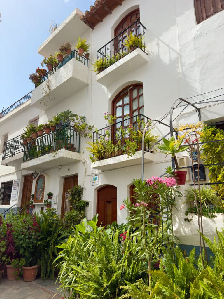 A white Spanish-style building with wooden doors and windows, balconies adorned with potted plants and flowers, and lush greenery in front—perfect for those enjoying scenic day trips from Granada under bright daylight.
