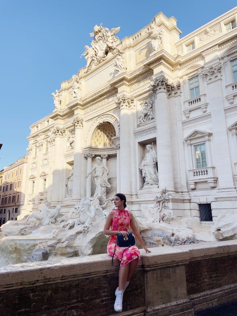 squares in rome: piazza di trevi. girl in pink dress sitting infront of white marble trevi fountain