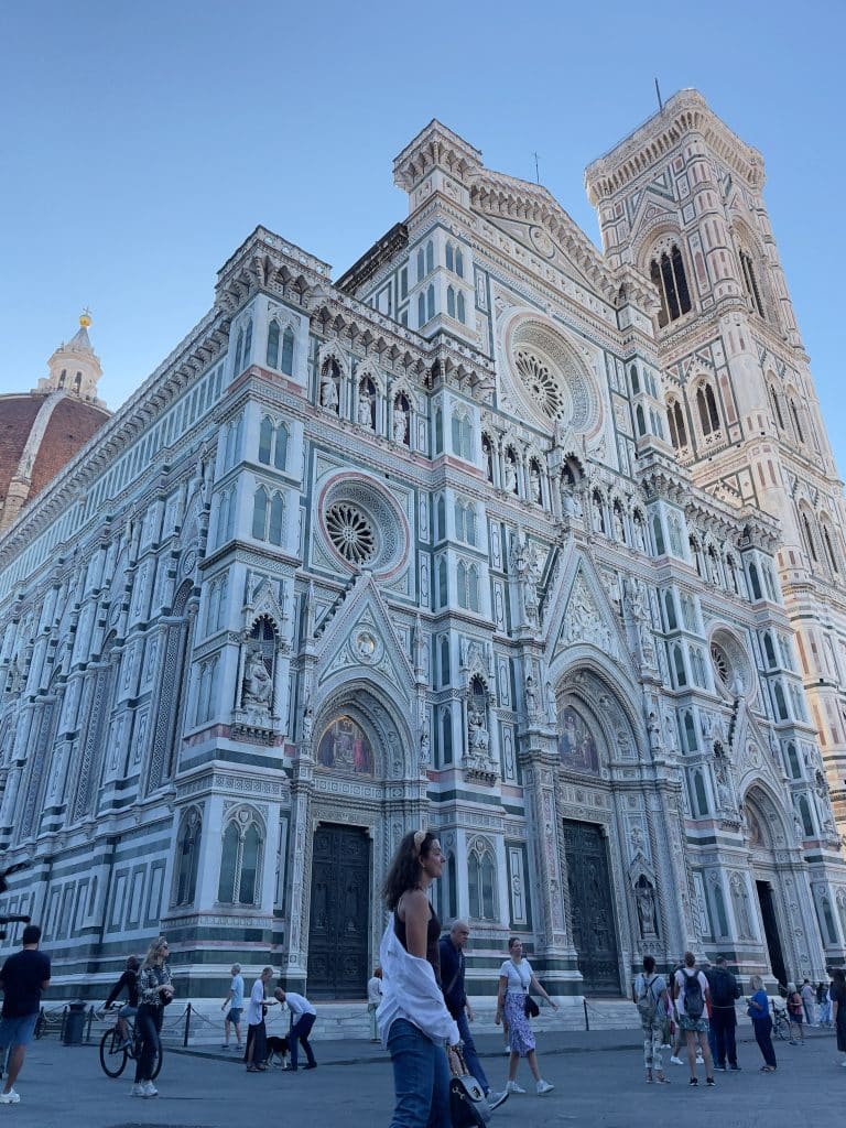 People walk in front of the ornate facade of Florence Cathedral, a must-see on Florence day trip from Rome, featuring intricate marble designs, sculptures, and the tall bell tower under a clear blue sky.