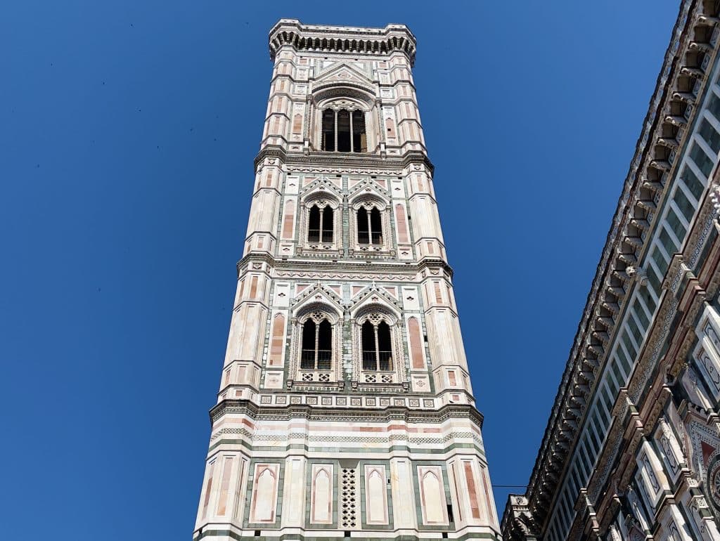 A tall, ornate Giotto bell tower with intricate architectural details and pointed arches rises against a clear blue sky. Much like the iconic Florence Duomo, the structure is adorned with decorative patterns in shades of white, pink, and green.
