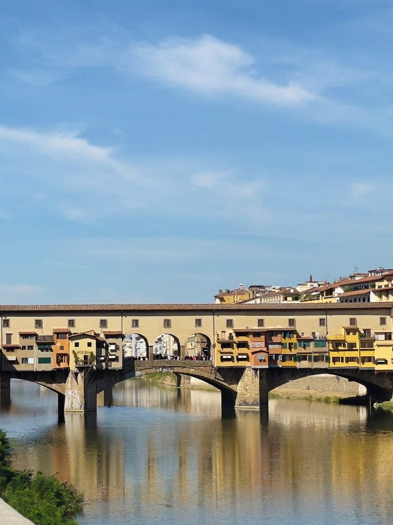 A historic arched stone bridge, Ponte Vecchio in Florence, with colorful buildings on top spans a calm river under a blue sky with wispy clouds; Florence, Italy is one of the best day trips from Rome.