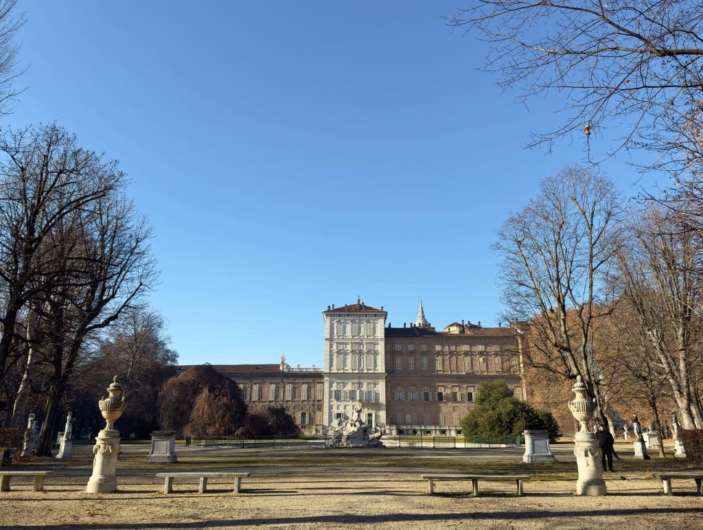 A grand historic palace with classical architecture stands at the end of a gravel path in a park flanked by leafless trees, stone benches, and sculptures under a clear blue sky. Visiting the Giardini Reali is one of the best free things to do in Turin.