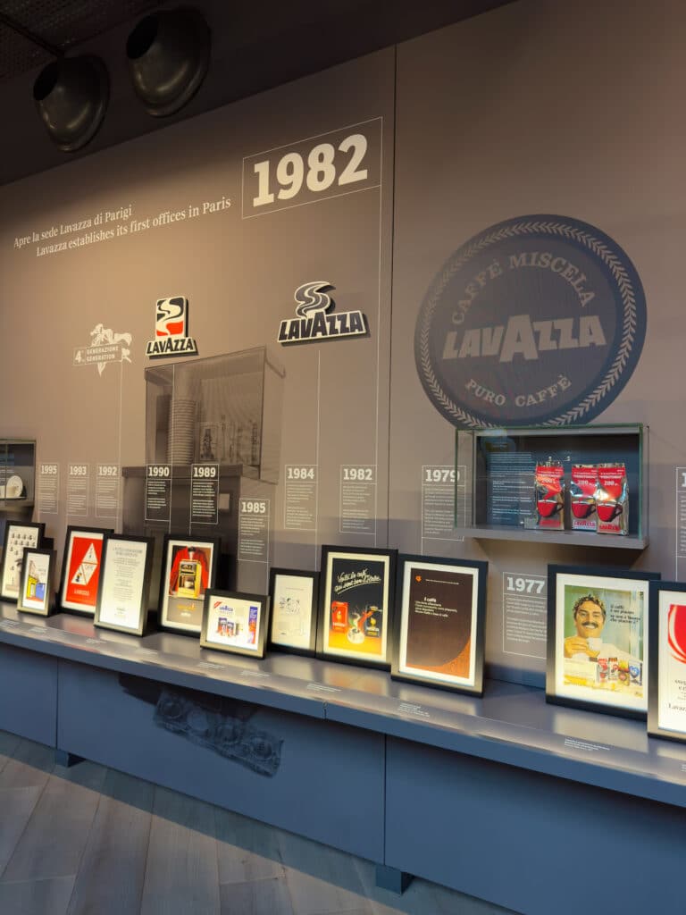 A display wall shows Lavazza’s history, featuring framed awards, vintage ads, coffee packages, and a large Lavazza logo—a reminder that Turin innovative heritage and why its worth visiting for coffee lovers. The timeline highlights 1982 when Lavazza established offices in Paris.