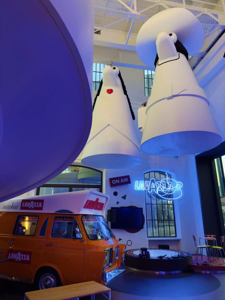 Large whimsical sculptures resembling a chess king and queen hang from the ceiling in the Lavazza Museum. Below, an orange Lavazza van, neon signs, and modern furniture create a vibrant, playful indoor scene. 
