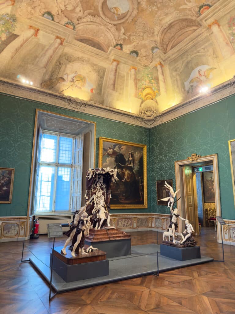 An ornate museum room in the Royal Residences of the House of Savoy, with green patterned walls, grand paintings, and a decorated ceiling. Modern abstract sculptures stand on pedestals—a must visit in Turin for art and history lovers.