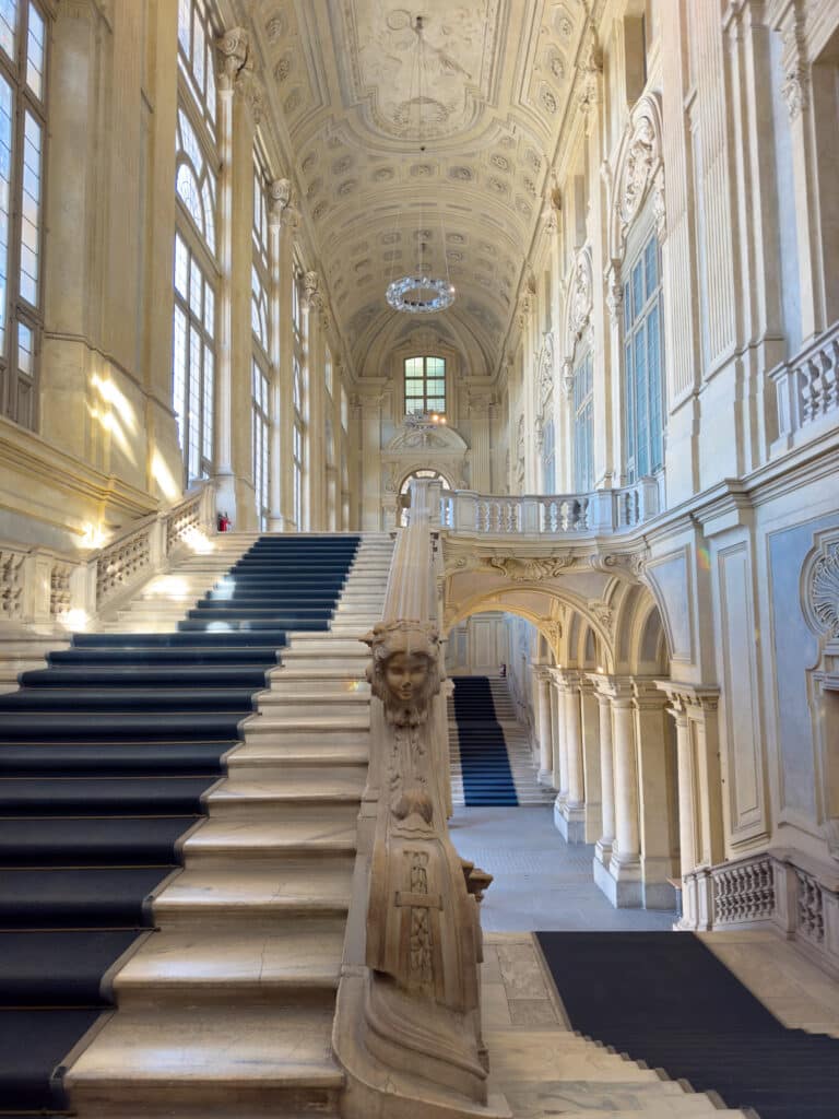 Grand staircase in an ornate, high-ceilinged hall—part of the Royal Residences of the House of Savoy. Sunlight streams through tall arched windows, highlighting decorative molding and chandeliers. A must visit in Turin for architecture lovers.