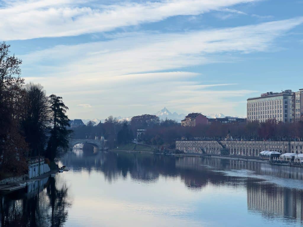 A calm river reflects trees and buildings on both sides, with a distant arched bridge and snow-capped mountains under a partly cloudy blue sky.