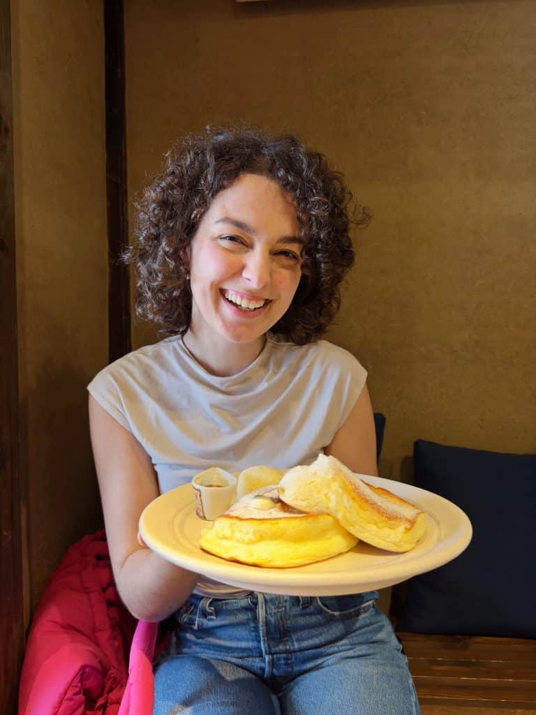 A smiling young woman with curly hair holds a plate of fluffy pancakes with a small container of syrup on the side. She is sitting in a cozy restaurant with wooden walls. Eating Japanese souffle pancakes at Cafe Tamon is one of the many best things to do in Kanazawa.