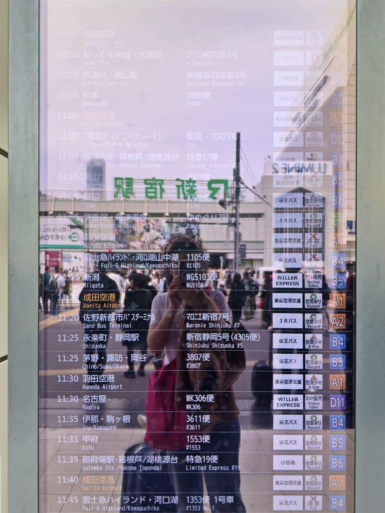 A reflective electronic timetable displays multiple trains' departure and arrival times in Japanese and English. In the background, a person takes a photo, capturing their reflection and a building with signs in Japanese.