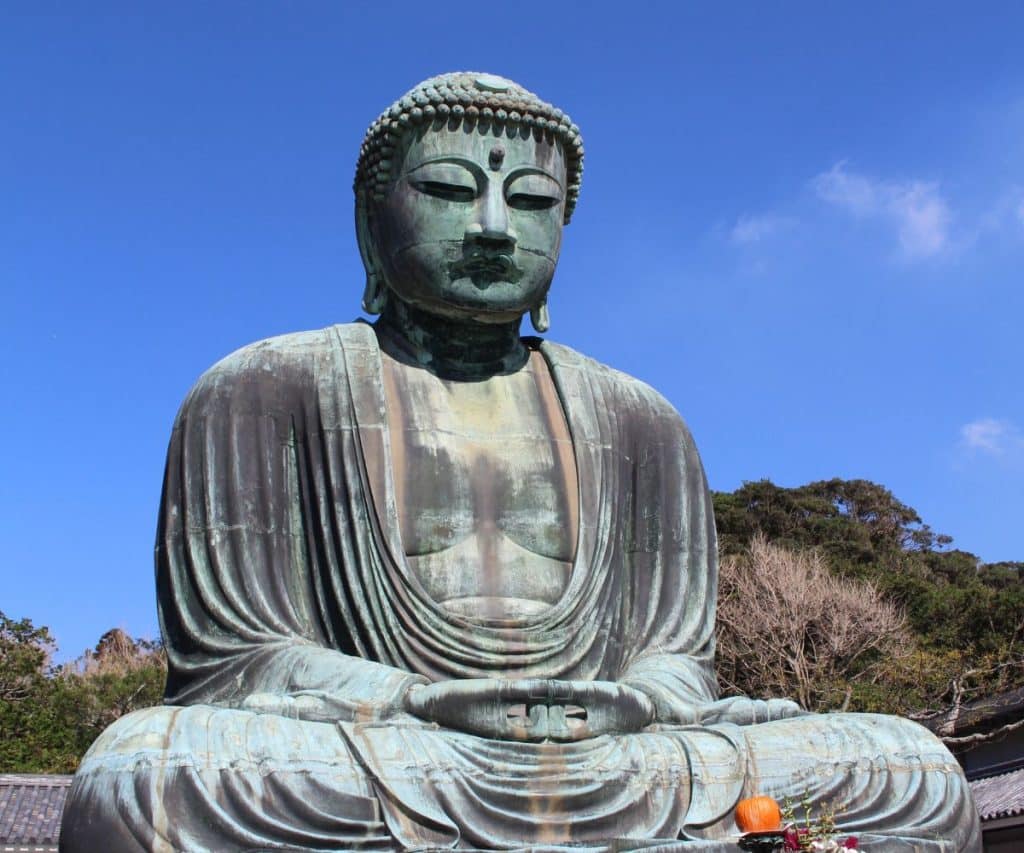 A large bronze statue of the Great Buddha, seated in a meditation pose with eyes closed, is set against a backdrop of trees and a clear blue sky. The statue has a serene expression and is adorned with intricate details, including a ushnisha on the top of its head. Kamakura is one of the best day trips from Tokyo.