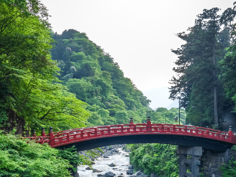 Shinkyo Bridge, a vibrant red arched bridge spans over a rocky stream in a lush, green forested area. Tall trees and dense foliage surround the bridge, with hills rising in the background under a partially clear sky. The scene is serene and picturesque. Nikko is one of the best day trips from Tokyo.