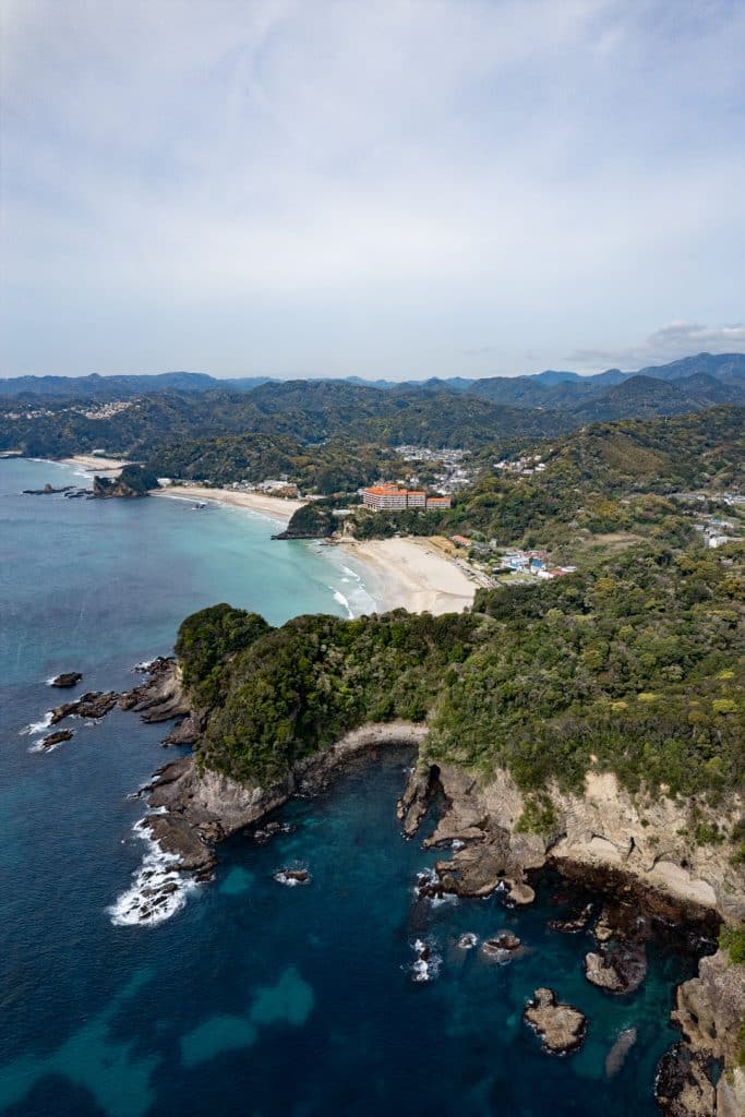 Aerial view of Shimoda, Japan, a picturesque coastal city and one of the best day trips from Tokyo. The scene features a sandy beach surrounded by lush green hills. The coastline includes rocky cliffs and clear turquoise waters, with buildings visible in the distance under a partly cloudy sky.