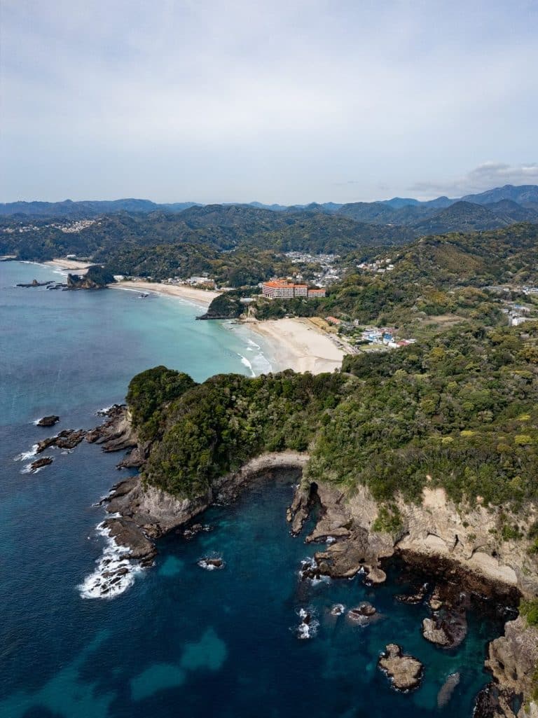 Aerial view of Shimoda, Japan, a picturesque coastal city and one of the best day trips from Tokyo. The scene features a sandy beach surrounded by lush green hills. The coastline includes rocky cliffs and clear turquoise waters, with buildings visible in the distance under a partly cloudy sky.