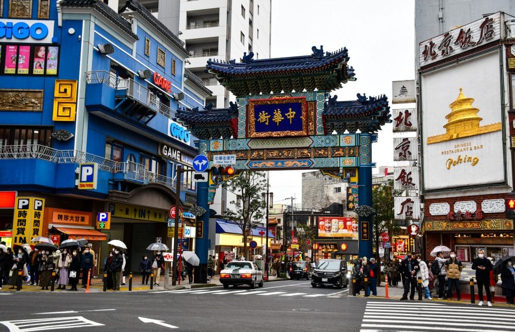 Street view in a vibrant urban area with people walking and cars driving. An intricate, colorful Chinese archway surrounded by buildings adorned with bright signs in various languages takes center stage. Yokohama is one of the best day trips from Tokyo.