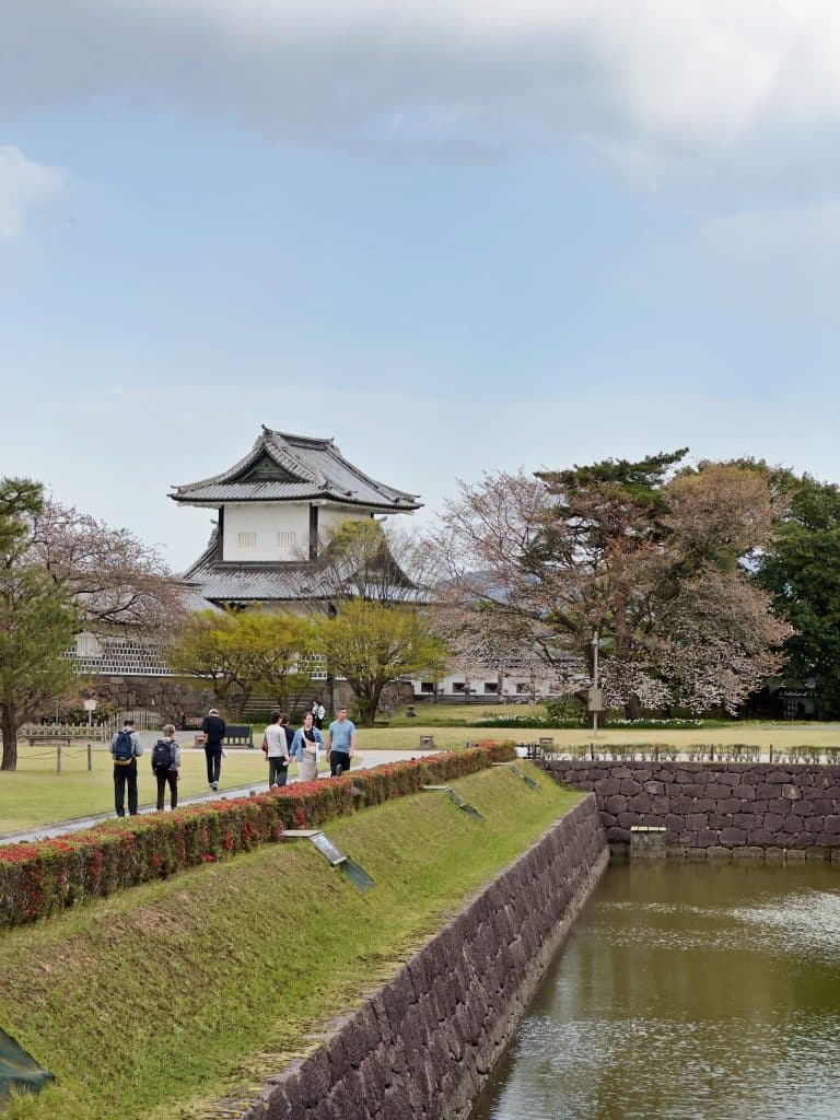 A serene landscape showcasing a traditional Japanese castle with a white facade and tiled roof. The castle is surrounded by trees, a stone wall, and a reflective moat. Visitors walk along a path by the moat, enjoying the scenery under a partly cloudy sky.