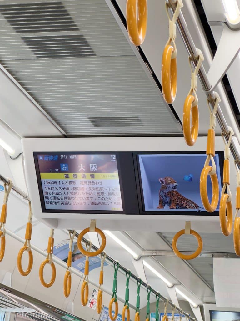 The interior of a train in Japan showing ceiling hand grips and a digital display. The display lists "Osaka" in large text, destination details in Japanese, and features an image of a leopard. The train is well-lit and has a clean, modern appearance.
