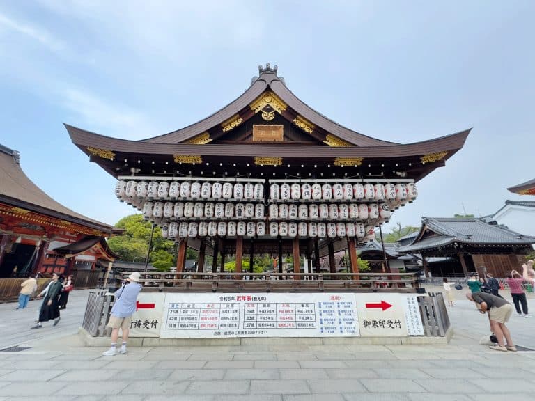 A traditional Japanese shrine with an elaborate wooden structure stands prominently. Many paper lanterns hang from the front, and various signs and boards are displayed below. People are walking and taking photos nearby. The sky above is clear and blue.