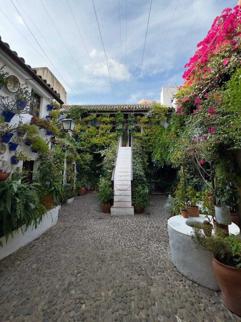 A picturesque courtyard with a cobblestone path, surrounded by white buildings adorned with lush green plants and vibrant pink flowers. A white staircase leads up to a balcony, and the sky above is partly cloudy.