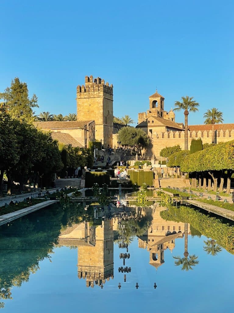 A sunlit castle with a tall tower and palm trees is reflected in a calm, rectangular pond. On your one day in Cordoba, the clear blue sky enhances the serene atmosphere of this historic site, surrounded by lush greenery.