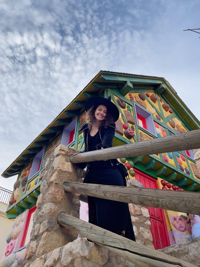 A person wearing a black hat and dress smiles while leaning against a wooden railing. Behind them is a colorful building with vibrant, decorative patterns and red doors. The sky is partly cloudy.